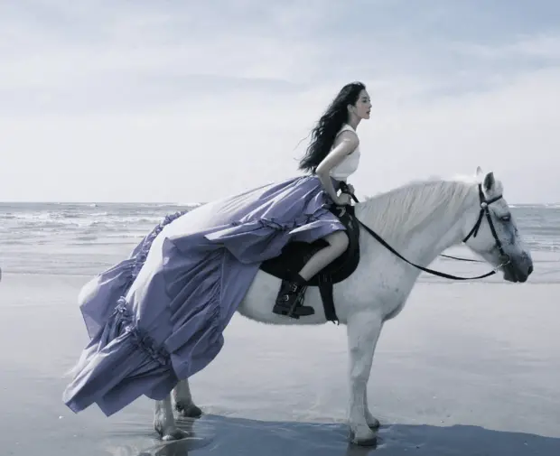 杨幂骑乘白马立于海边,紫纱裙摆随风轻扬。