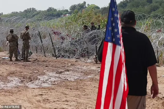 ▎一名男子举着一面旗帜，看着德克萨斯州国民警卫队成员与一群移民对峙。图源：每日邮报